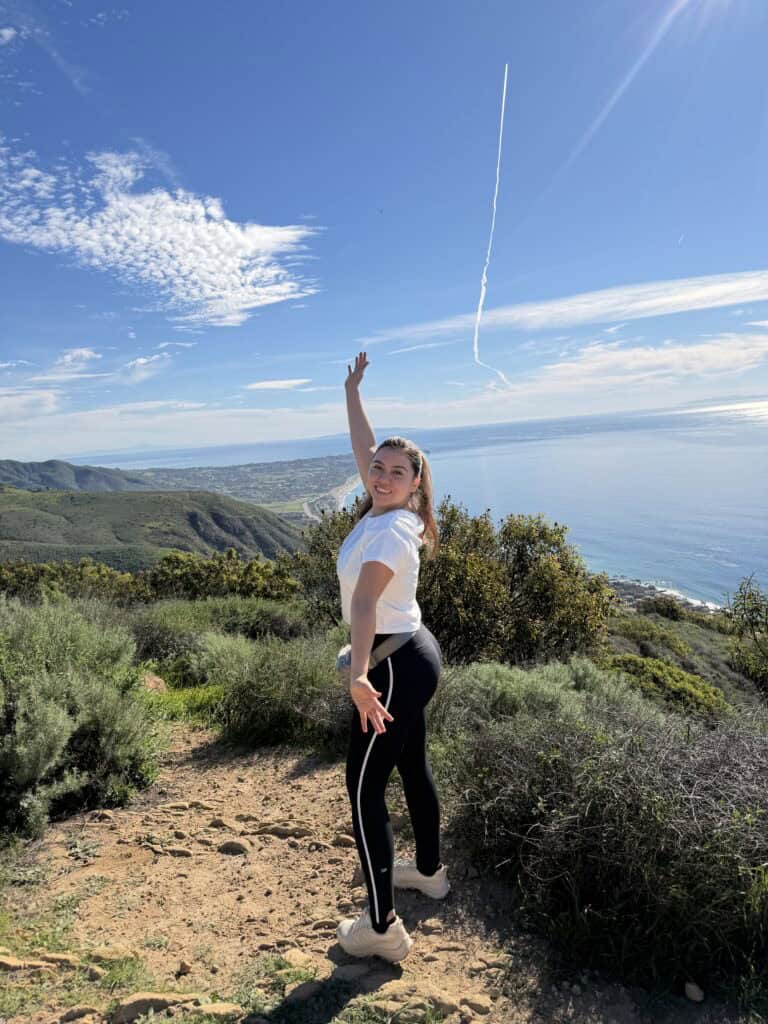 Founder of layers of beauty in workout clothes hiking a coastal trail in Los Angeles with her arm raised, overlooking the Pacific Ocean – representing daily movement as part of a time management routine
