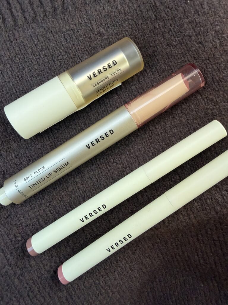 Four Versed Skincare lip products laid on a dark textured surface including two Lip Frame Defining Lip Liners, the Soft Gloss Tinted Lip Serum, and the Cashmere Color Smoothing Lip Blush