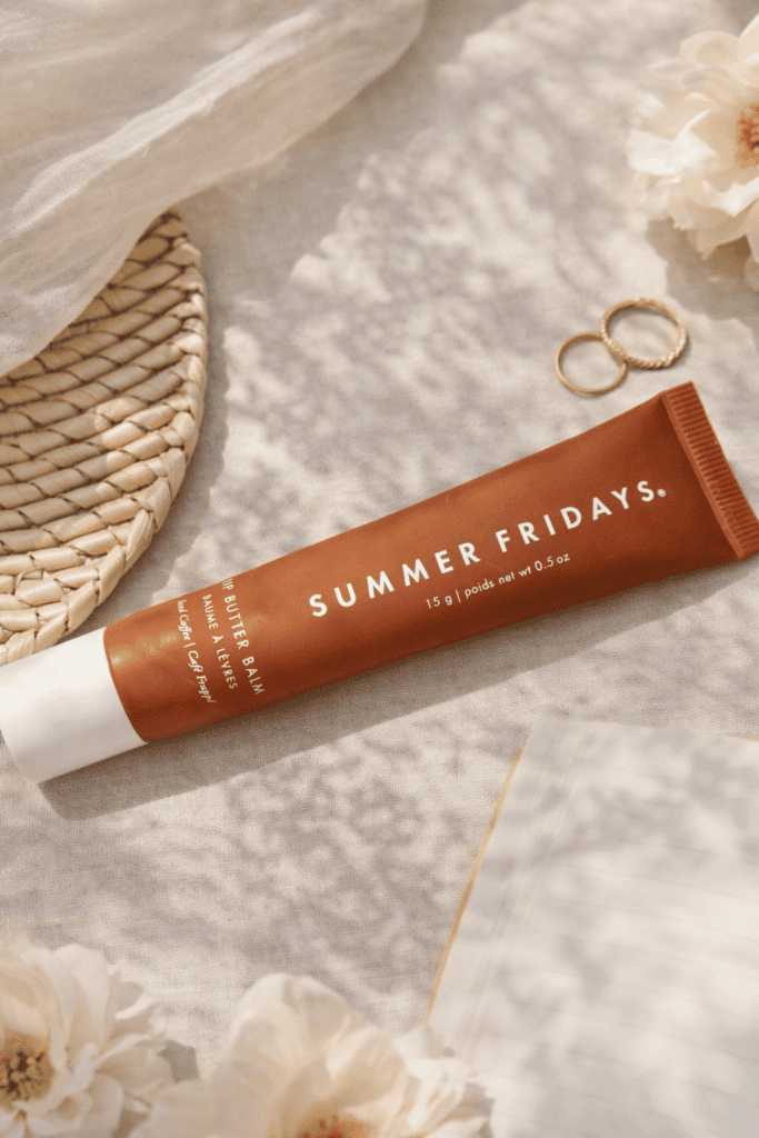 Summer Fridays Lip Butter Balm tube in Iced Coffee shade laying flat on a cream linen surface surrounded by white flowers and gold rings