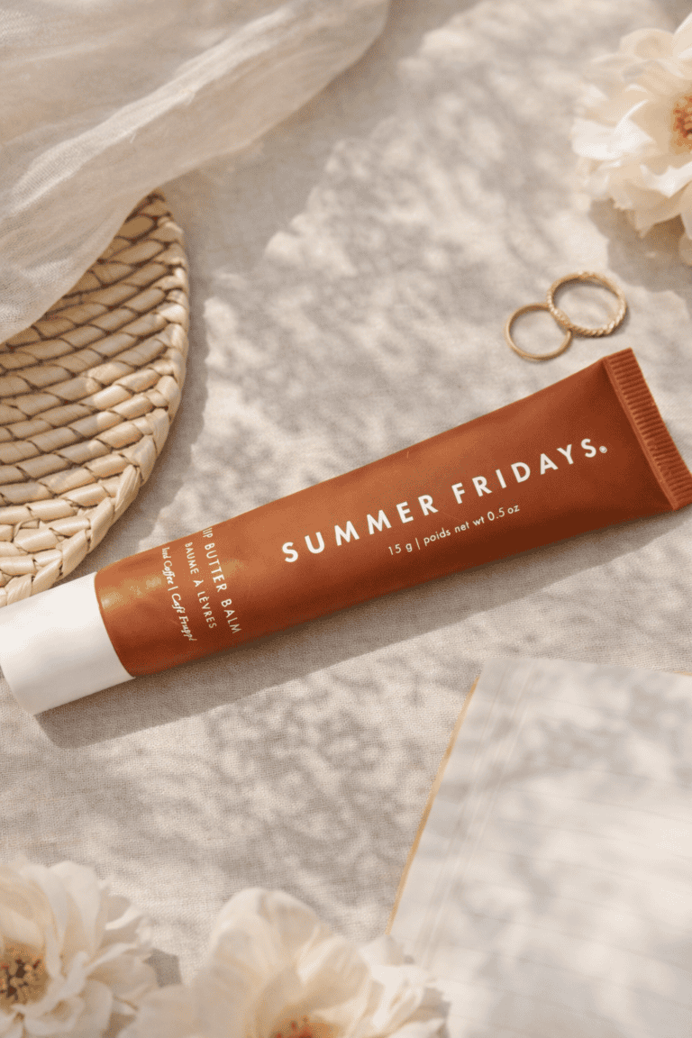 Summer Fridays Lip Butter Balm tube in Iced Coffee shade laying flat on a cream linen surface surrounded by white flowers and gold rings