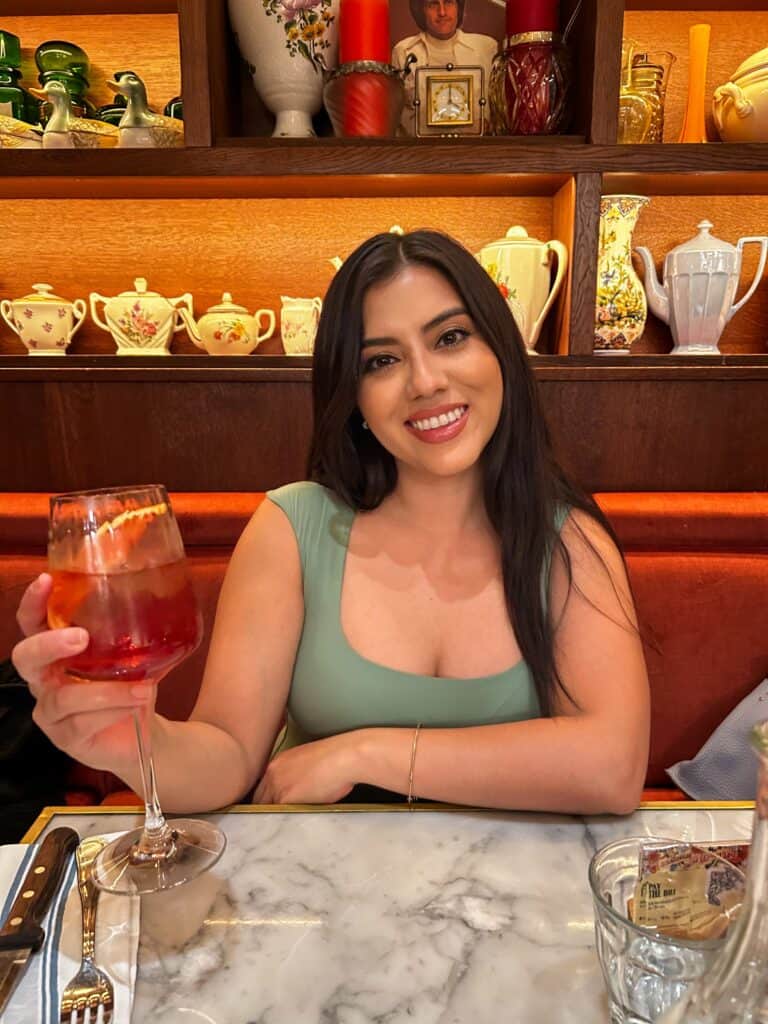 Jasmine Del Toro sitting at a marble table at a restaurant in Los Angeles, holding a wine glass, wearing a sage green sleeveless dress – date night outfit inspiration